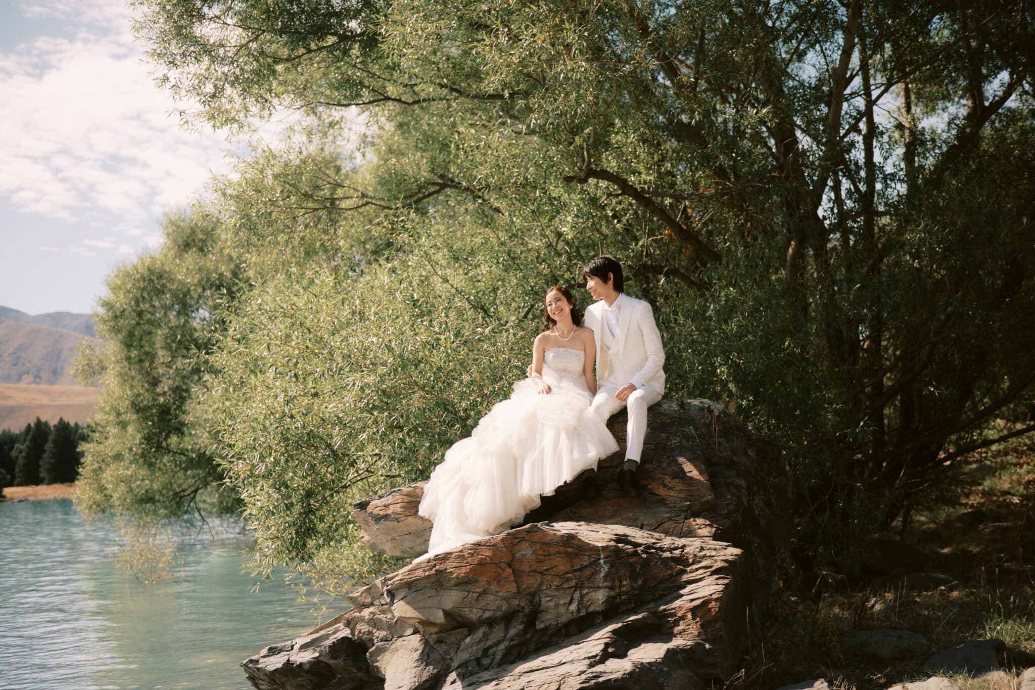 Queenstown Tekapo New Zealand Heli Wedding Elopement Pre-Wedding Shoot Photographer クイーンズタウン　テカポ　ニュージーランド　エロープメント 前撮り　フォトウェディング　結婚式 | A couple in wedding attire seated on a rocky outcrop by the serene Lake Tekapo, surrounded by greenery for their pre-wedding shoot.